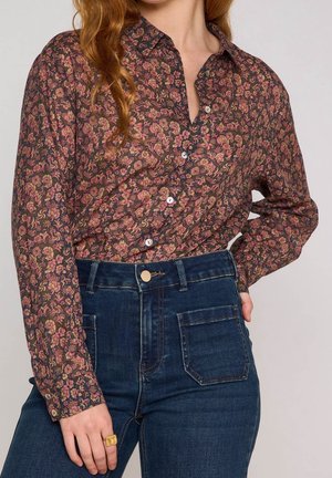 Tenue féminine avec une chemise à manches longues boutonnée à motif floral foncé rentrée dans un jean en denim bleu taille haute, accessoirisée d’une bague en or.