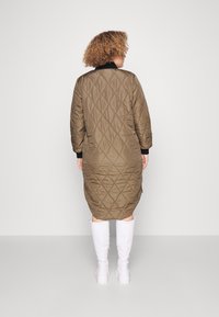 Persona con cabello rizado que lleva un abrigo acolchado verde oliva hasta la rodilla y botas blancas hasta la rodilla, de pie de espaldas sobre un fondo liso.