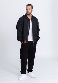 Schwarze Bomberjacke mit Reißverschluss vorne und gerippten Bündchen, kombiniert mit schwarzen Cord-Hose im Cargo-Stil und weißen Sneakers. Sichtbares Detail mit orangefarbigem Etikett.