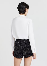 Camicia bianca a maniche lunghe con dettagli a bottoni sul colletto, abbinata a pantaloncini corti neri con paillettes caratterizzati da una superficie testurizzata e un design aderente.