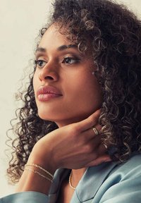 Schmuck in Goldfarbe umfasst Ringe und Armbänder. Das Modell hat lockiges, schulterlanges Haar und trägt ein hellblaues Kleidungsstück mit glatter Textur.