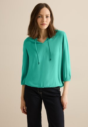 Une blouse turquoise à manches courtes avec un col en V et un cordon de serrage. Fabriquée dans un matériau léger, elle présente une texture lisse et une coupe décontractée.