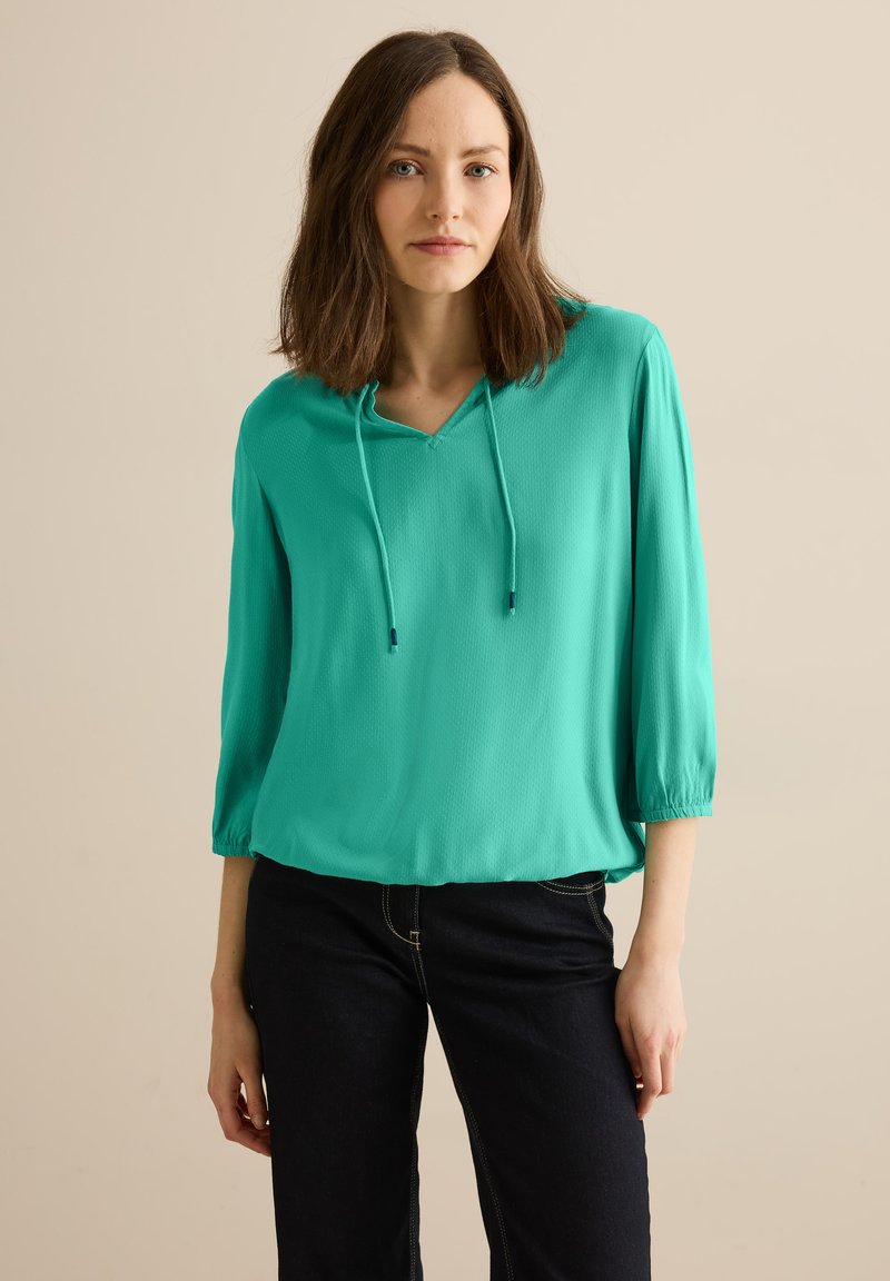 Una blusa turchese a maniche corte con scollo a V e coulisse. Realizzata in materiale leggero, presenta una texture liscia e una vestibilità comoda.