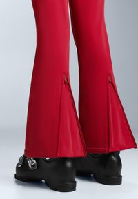 Piros, bővülő szárú nadrág cipzáras díszítéssel a szegélyén, sima anyagból, fekete, stílusos lábbelivel párosítva, melyen fémes kiegészítők találhatók.