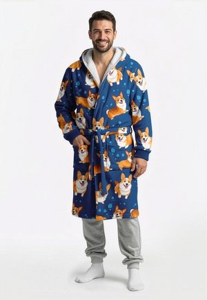 Usmievajúci sa muž stojaci, oblečený v modrom flísovom župane s motívmi psov corgi, sivé tepláky a biele ponožky na jednoduchom pozadí.