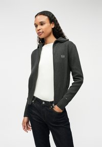 Grå hettejakke med ribbestrikkede mansjetter og krage, med et lite logo på venstre bryst. Kombinert med mørk denimjeans.