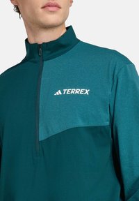 Tyrkysová polovičná zipová mikina, vyrobená z ľahkej tkaniny. Obsahuje logo Terrex v bielej farbe na hrudi a kontrastnú textúru na pleciach.