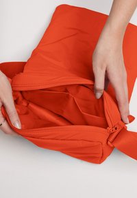 Borsa arancione imbottita con una texture liscia, dotata di una zip e di una tasca interna. Il tessuto sembra leggero e resistente.