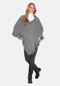Frau mit langen blonden Haaren, die einen grauen poncho mit Fransen, einen weißen Rollkragenpullover, dunkle Jeans und schwarze kniehohe Stiefel trägt und mit überkreuzten Beinen steht.