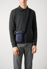 Mornarska crossbody torba z zadrgo na sprednjem žepu, okrašena z črtastim vzorcem. Narejena iz teksturiranega materiala, pritrjena na nastavljiv trak.