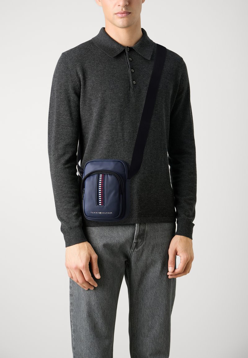 Mornarska crossbody torba z zadrgo na sprednjem žepu, okrašena z črtastim vzorcem. Narejena iz teksturiranega materiala, pritrjena na nastavljiv trak.