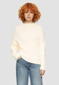 Cremefarbener gerippter Strickpullover mit hohem Kragen und lockerer Passform, kombiniert mit hellblauen Jeans. Er hat lange Ärmel und einen subtil asymmetrischen Saum.