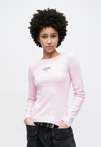 T-shirt de malha rosa claro de manga comprida com o logótipo "GUESS" em letras escuras, combinada com calças pretas de vários bolsos e um cinto com tachas.