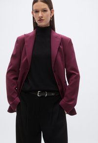 Bordeauxrode blazer met revers; zwarte coltrui eronder; zwarte broek met hoge taille; riem met zilveren gesp; gladde stof met een getailleerde pasvorm.