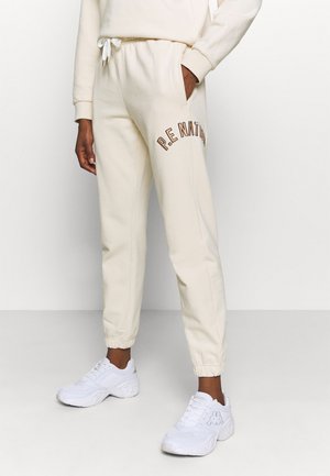 Beige sweatpants met een trekkoord in de taille, elastische boorden en "P.E. NATION" in bruin geborduurd. Gecombineerd met witte sport schoenen.