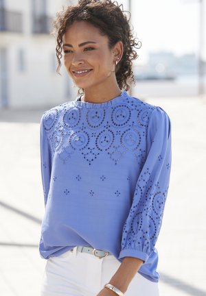 Light blue blouse with eyelet embroidery featuring circular and floral patterns. Rounded neckline, long sleeves with gathered cuffs, and soft texture.