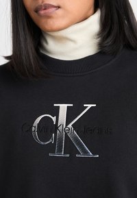 Svart sweatshirt med ribbad krage, med en silverfärgad "CK"-logotyp och broderad "Calvin Klein Jeans" text nedan i svart.