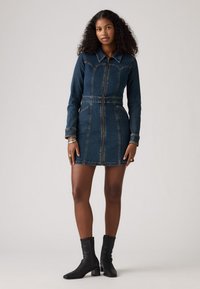 Abito in denim scuro con maniche lunghe, vita aderente e zip frontale. Presenta un colletto a punta e dettagli con polsini. Indossato con stivaletti neri con tacco.