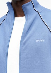 Κοντινό πλάνο ενός ατόμου που φοράει ανοιχτό μπλε μπουφάν με φερμουάρ και λευκές ραφές, με το λογότυπο "BOSS" κεντημένο στο στήθος πάνω από λευκό πουκάμισο.