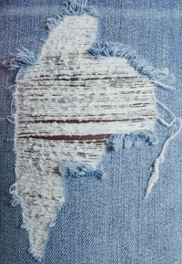 Jasnoniebieski dżins z dużym, nieregularnie kształtowanym przetarciem z postrzępionymi brzegami i zniszczoną teksturą.