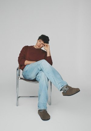 Hnědý svetr, světle modré džíny, šedo-hnědé nazouváky, sedící na chromované židli na šedém pozadí. Neformální outfit s uvolněným postojem.