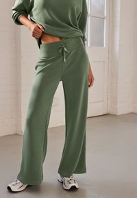 Green wide-leg sweatpants with an elastic waistband and drawstring detail, made from soft fabric; paired with gray athletic shoes.