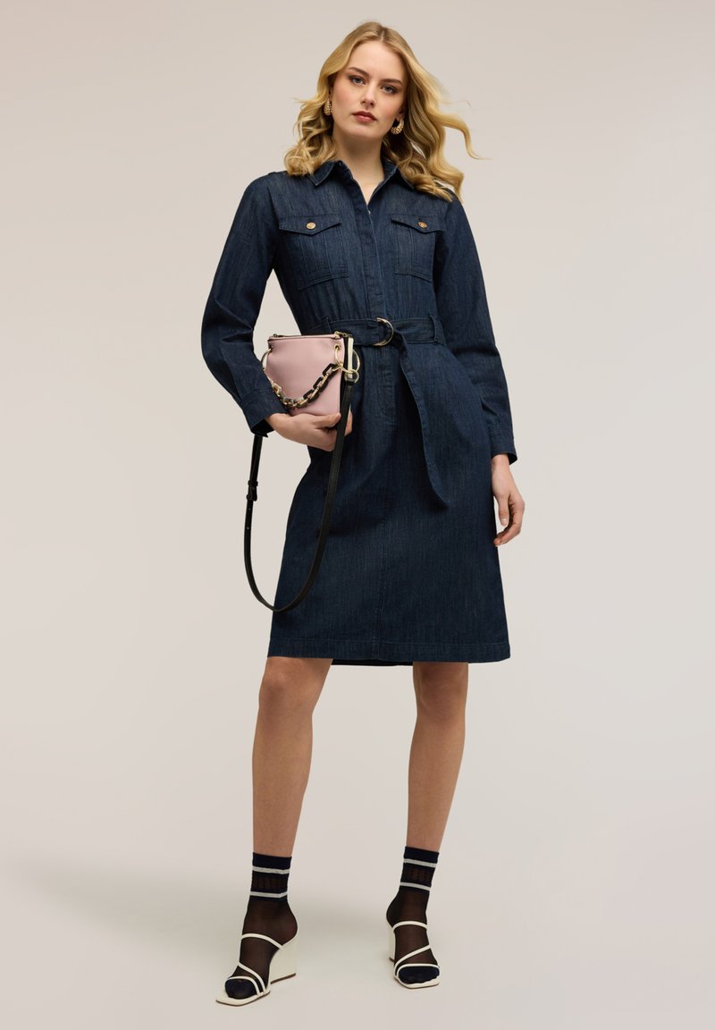 Femeie blondă purtând o rochie din denim închis la culoare, cu curea, șosete negre transparente, sandale albe, ținând o geantă mică roz cu o curea aurie din lanț.