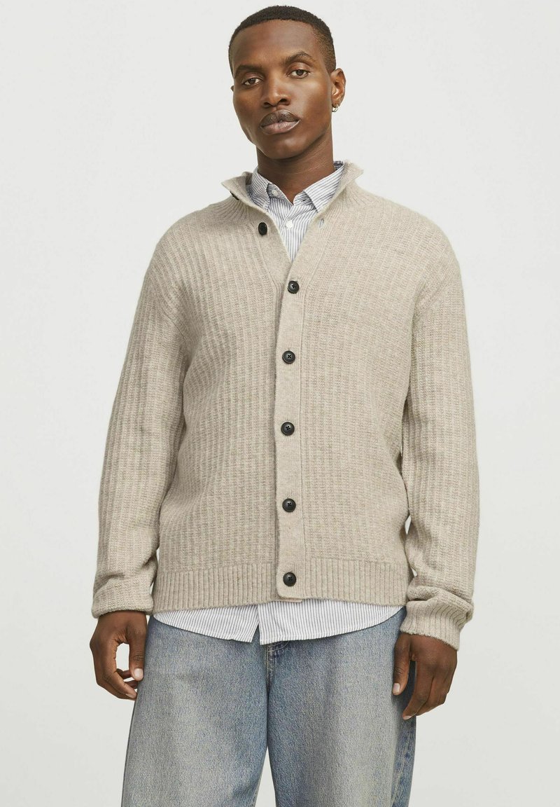 Beige gestrickte Strickjacke mit V-Ausschnitt und gerippten Bündchen. Besitzt schwarze Knöpfe und eine strukturierte Oberfläche, die über einem blau gestreiften Hemd getragen wird.