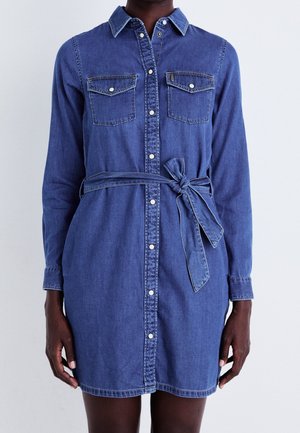 Kvinde iført en blå denimskjortekjole med lange ærmer, knapper foran, brystlommer og et bundet bælte i taljen.