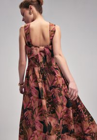 Blumiges Maxikleid mit einem tiefen Rückenausschnitt, versehen mit einer breiten rosa Schleife in der Taille. Der Stoff zeigt lebendige Pinktöne und erdige Farben in einem malerischen Muster.