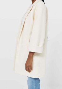 Cream-colored oversized coat with a lapel collar, rolled cuffs, side pockets, and a straight silhouette, made from a soft, textured fabric.