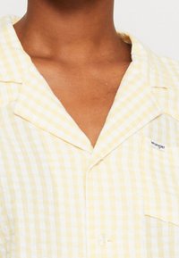 Chemise à carreaux jaunes et blancs en tissu léger, avec un col à encoches et une petite étiquette logo sur la poche.