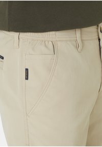 Beige trousers with a smooth texture, featuring a side pocket and a visible brand label. The waistband has elastic for comfort.