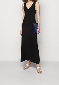Svart ärmlös maxiklänning med V-ringning, figurnära överdel och utställd nederdel. Kombinerad med vita sandaler med remmar och en blå clutch.