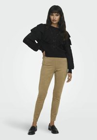 Pull en tricot noir avec des accents froufroutants, associé à un pantalon en daim beige. Le modèle porte des mocassins noirs. Design simple et moderne avec des détails texturés.