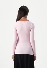 Rosa ribbad långärmad topp med scallopad halsringning och volang ärmar, bärs tillsammans med en svart plisserad kjol. Mjuk textur, figurnära design.
