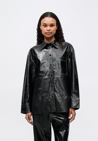 Kvinde iført en skinnende sort læderbutton-up skjorte med en brystlomme og matchende læderbukser, der står mod en enkel lys baggrund.