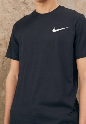 Person trägt ein schlichtes schwarzes Kurzarm-T-Shirt mit einem weißen Nike-Swoosh-Logo auf der Brust vor einem strukturierten Stein-Hintergrund.
