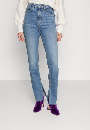 Femme portant un jean bleu taille haute avec des fentes devant les jambes et des bottines violettes à talons, associée à un pull blanc.