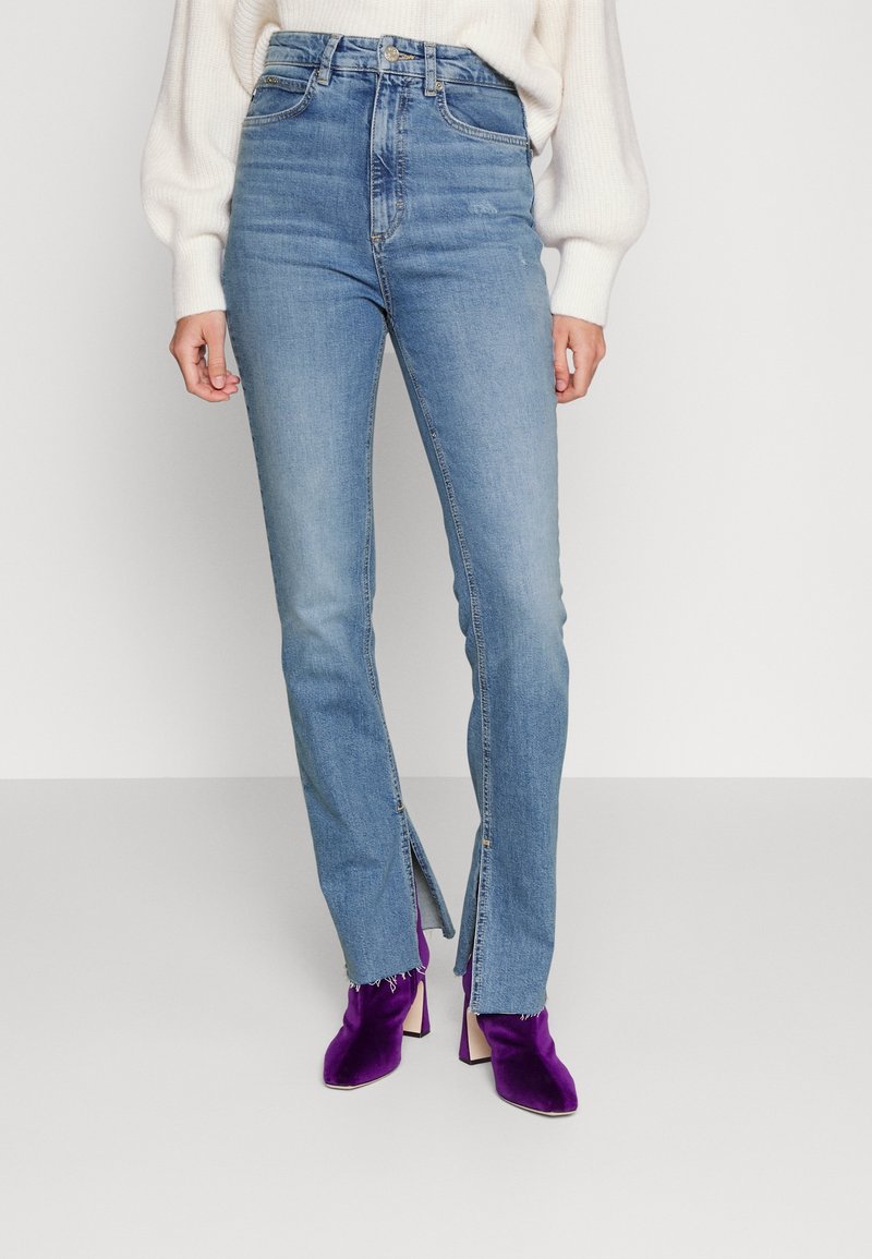 Femme portant un jean bleu taille haute avec des fentes devant les jambes et des bottines violettes à talons, associée à un pull blanc.