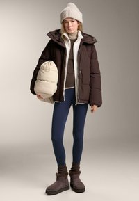 Smeđi puffer jakna, bijela unutarnja sloj, tamnoplave tajice, sivi čizmi i bež torba. Tekstuirana pletena kapa upotpunjuje outfit.