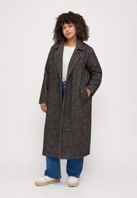 Lang, mørkebrun frakk med et subtilt herringbone-mønster, med store slag og dobbeltrad knapper, parret med avslappet denim og sko.