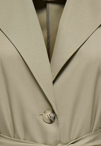 Gros plan sur un trench-coat beige avec de larges revers, un seul bouton en écaille de tortue fermé, et une ceinture en tissu assorti nouée à la taille.