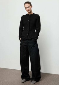 Czarny kardigan z fakturowanym wykończeniem, okrągłym dekoltem i zapięciem na guziki, w połączeniu z luźnymi czarnymi dżinsami i szarymi butami.