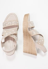 Sandales compensées beiges avec des brides croisées ornées de perles et de strass, sur une semelle plateforme tissée et texturée.