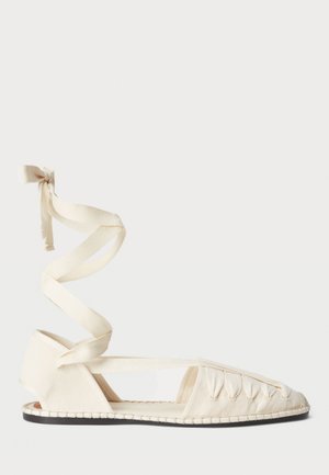 Kremowe płaskie buty z tkaniny z zaostrzonym noskiem, zapięciem w formie krzyżujących się pasków i teksturowaną powierzchnią, z czarną podeszwą i ozdobnym przeszyciem.