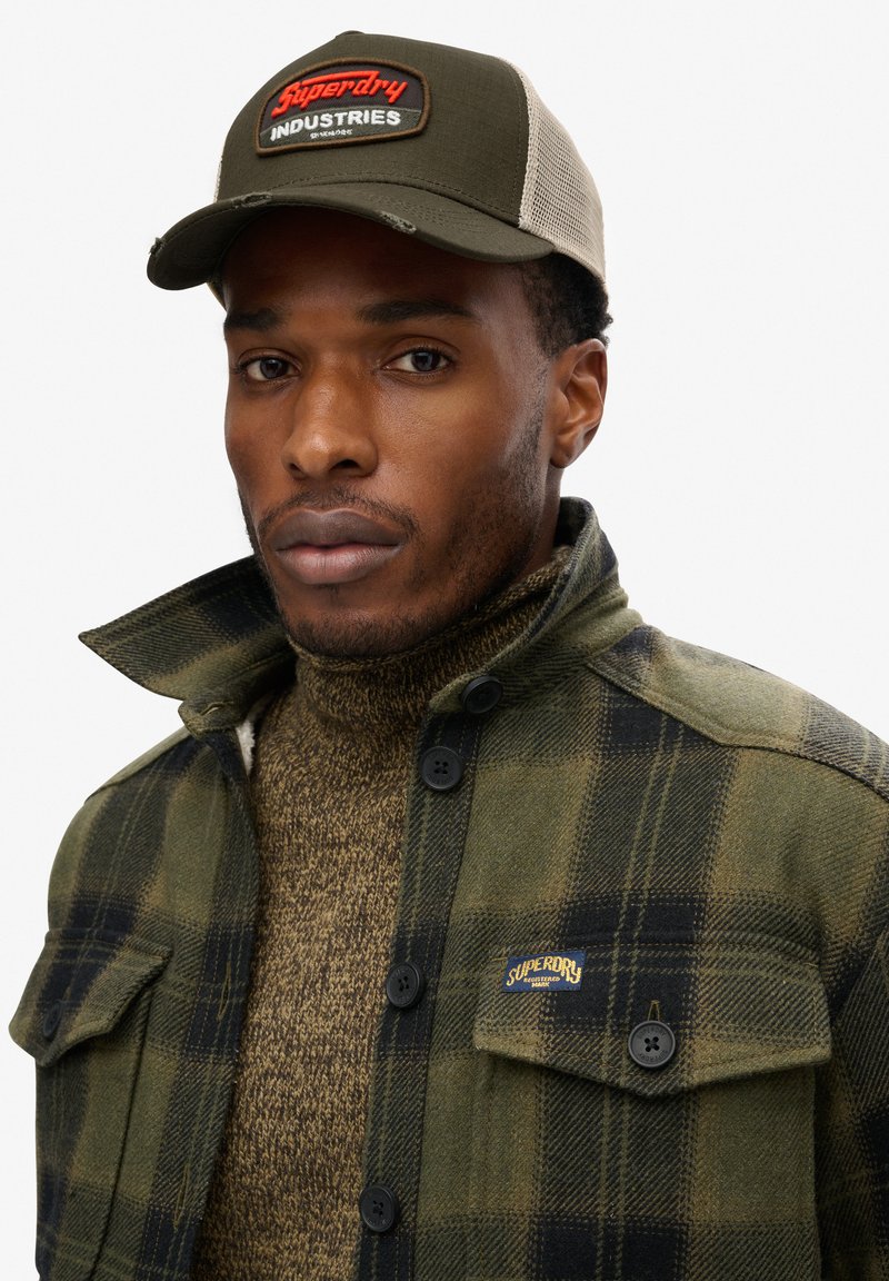 Chaqueta de cuadros verde con acentos negros, combinada con un suéter de cuello alto de punto marrón. La gorra es de color verde oliva con un parche de logo rojo.