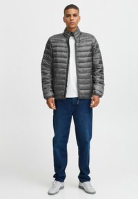 Grijze, gewatteerde pufferjack met een hoge kraag, voorkant rits en elastische manchetten, gedragen over een wit shirt en blauwe jeans.