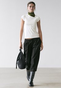 Vrouw draagt een witte top met korte mouwen, zwarte losse broek, zwarte laarzen, olijfgroene sjaal, en draagt een zwarte leren handtas terwijl ze binnen loopt.