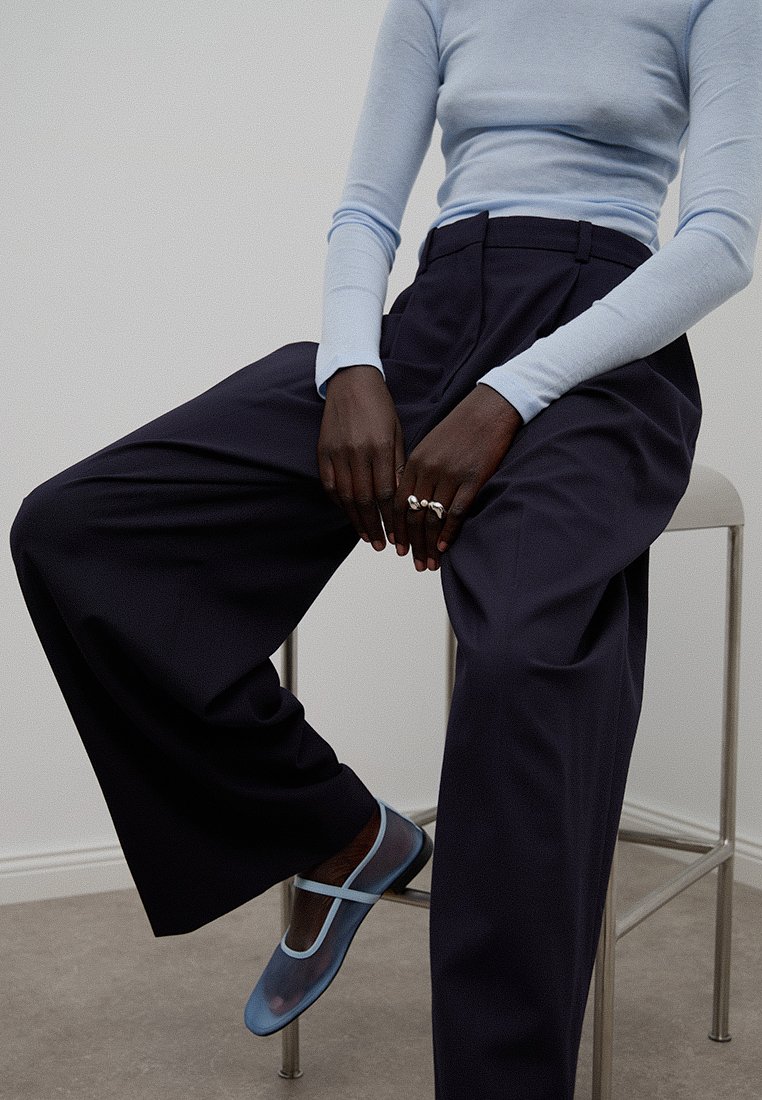 Jasnoniebieska bluzka z długim rękawem w połączeniu z ciemnymi szerokimi spodniami. Stopy ozdobione przezroczystymi niebieskimi balerinami, które mają gładką fakturę.